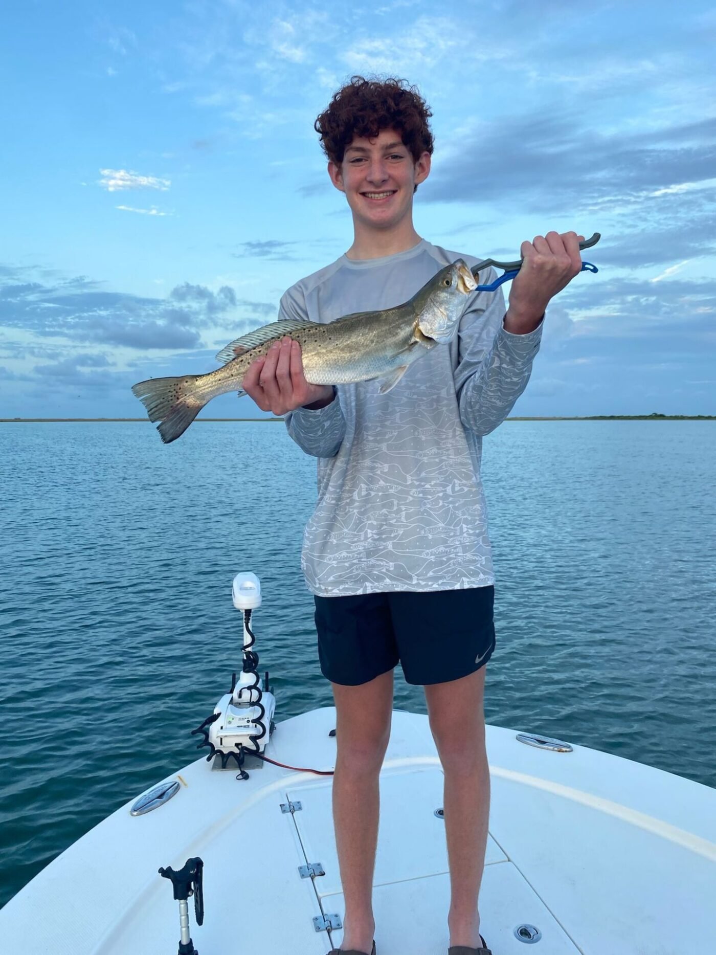 Teen boy proudly holding a freshly caught speckled trout on a fishing boat in calm coastal waters with blue skies