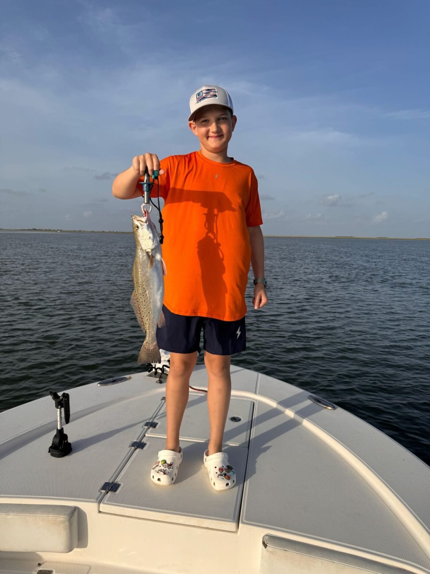 Smiling young boy in bright orange shirt proudly holding a speckled trout on a fishing boat during a Frenzy charter trip at sunrise
